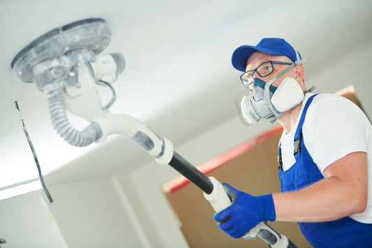 A man with a dust mask on sanding drywall with a pole sander